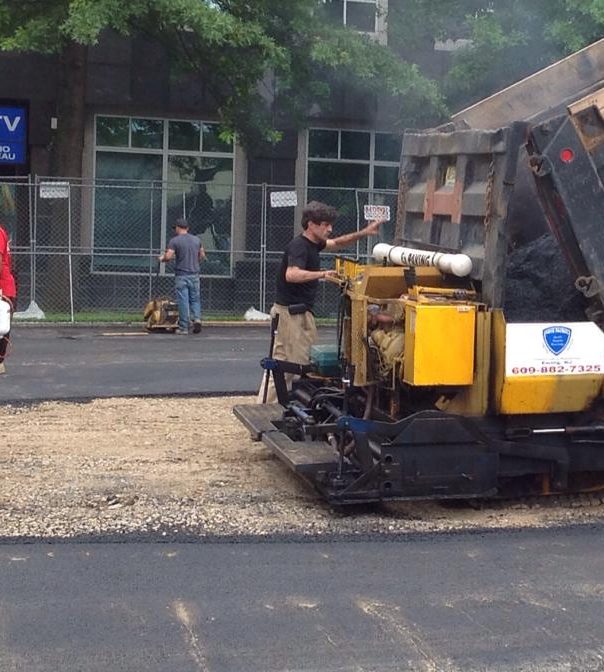 Worker operating a small asphalt paver over a gravel patch in front of a commercial building
