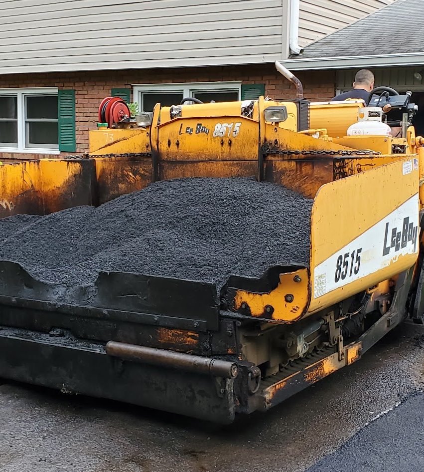 Close-up of a yellow asphalt paver hopper filled with hot mix asphalt in a residential driveway