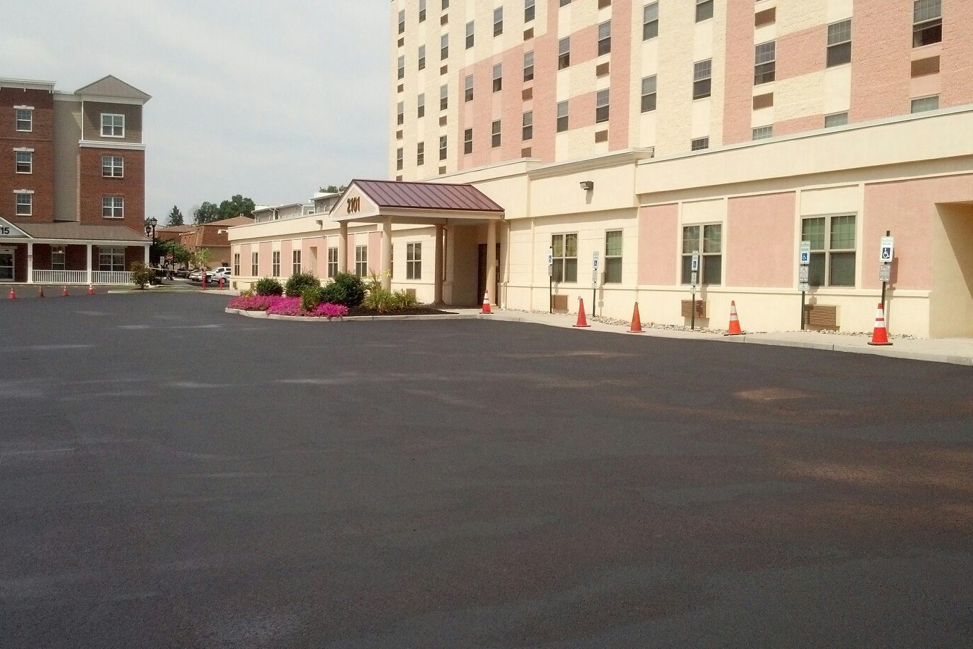 Freshly paved black asphalt parking lot in front of a large multi-story building, with traffic cones