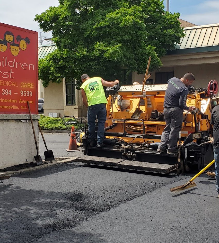 Paving crew operating a yellow asphalt paver beside a commercial building