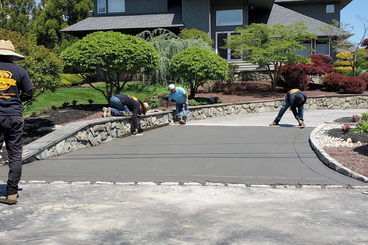 Crew finishing a new concrete driveway section bordered by stone curbing and landscaped beds