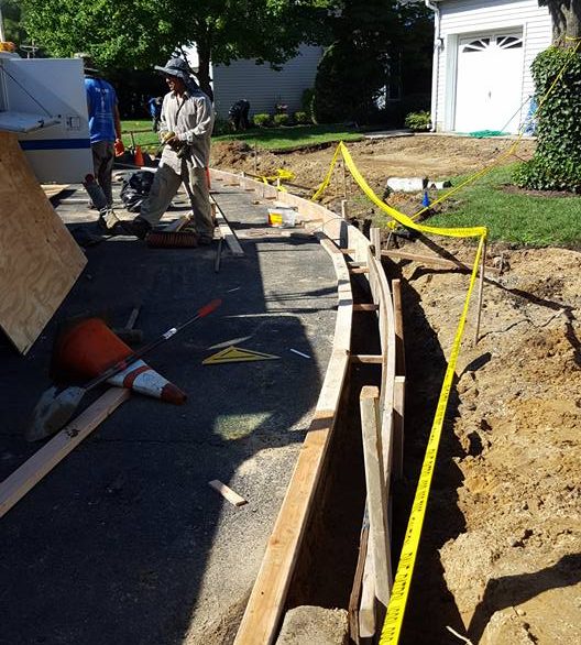 Excavated trench with wooden forms and a tape measure beside an asphalt driveway, worker in the background
