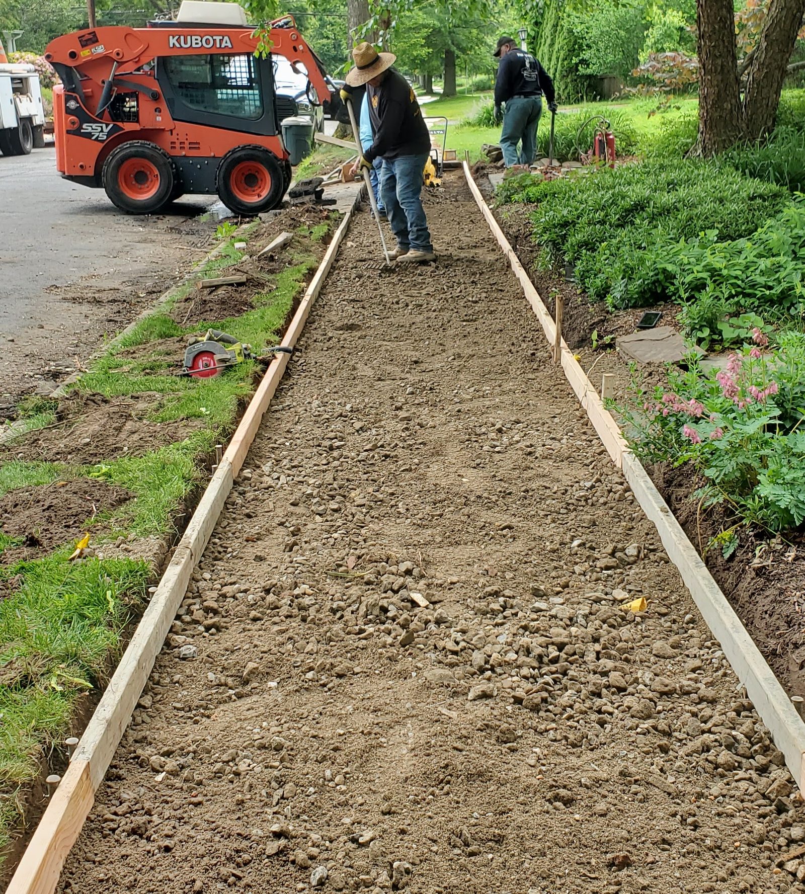 Crew grading a gravel base between wooden forms for a new walkway, with an orange skid steer nearby