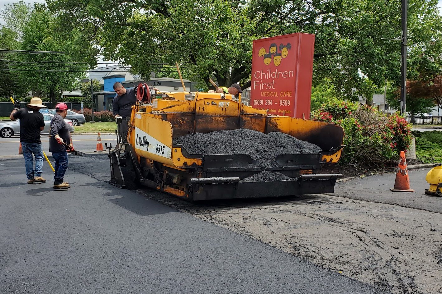 Crew with a yellow asphalt paver repairing pavement at a business entrance, traffic cones nearby