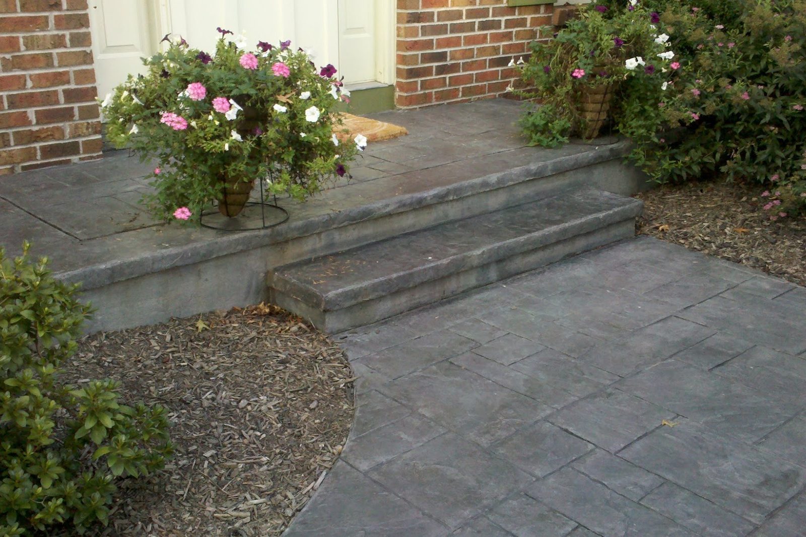Gray stamped concrete walkway and steps leading to a brick home entrance with flower pots