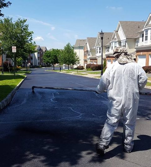 Technician in a white protective suit spraying sealcoat on an asphalt road lined with townhouses