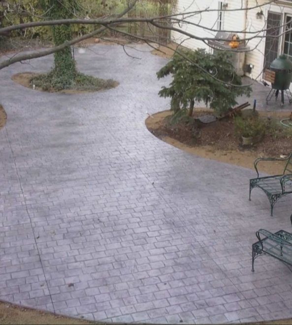 Large curved gray stamped concrete patio wrapping around a tree bed, with patio chairs