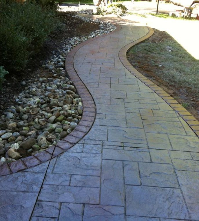 Curved gray stamped concrete walkway with a red brick-pattern border and river rock beds